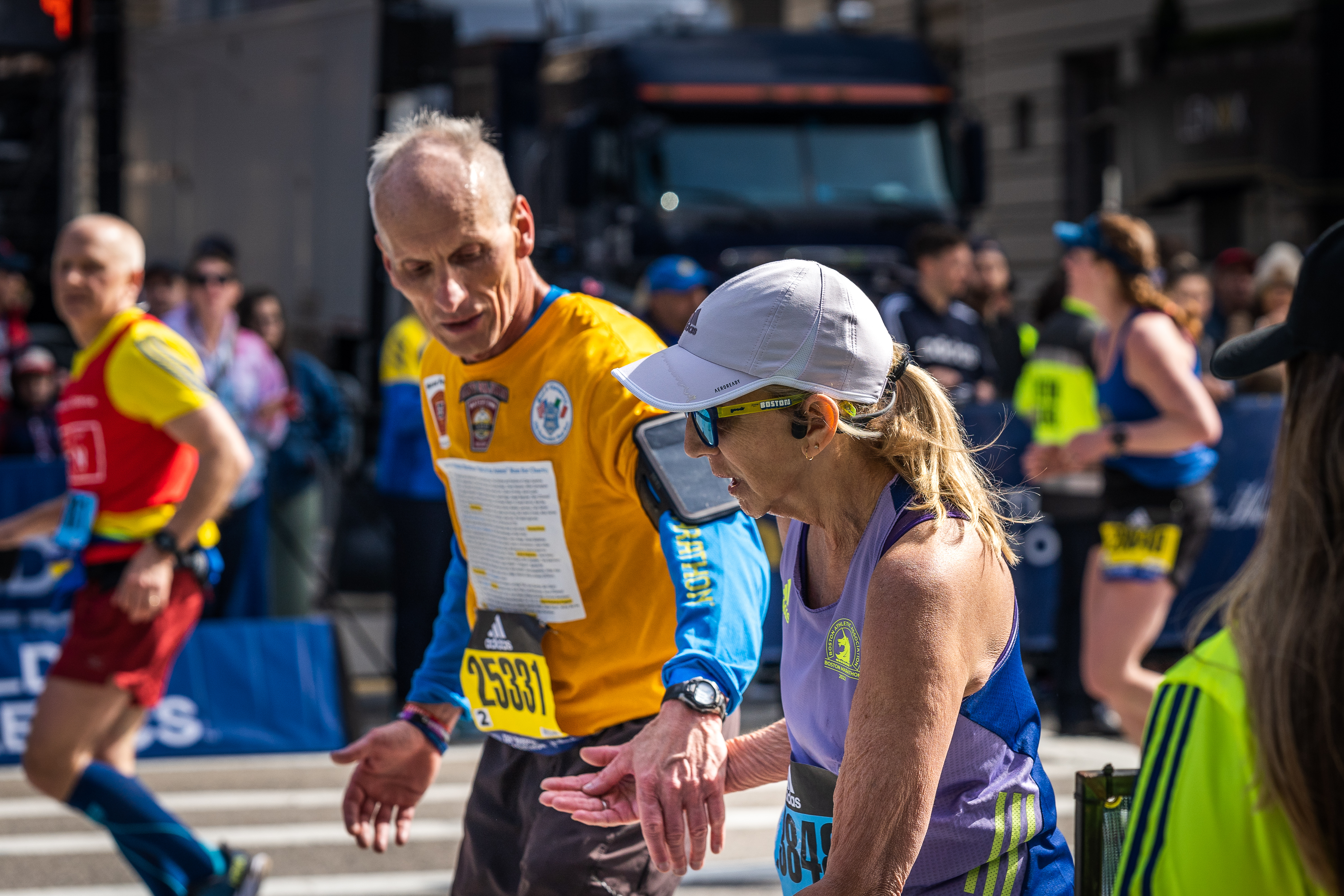An older man offering a hand to an older woman that is showing difficulties in completing the Boston Marathon